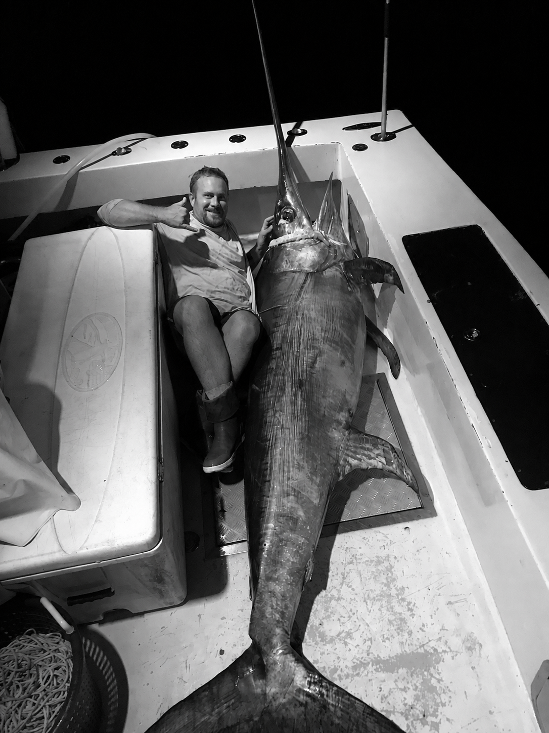 Fisherman seated beside a giant swordfish on the deck of an offshore boat at night in Florida, with the fish longer than he is tall.