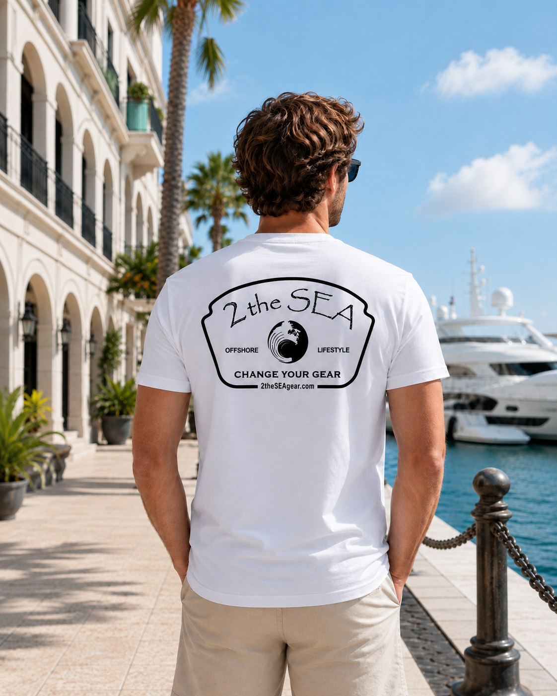Back view of a man wearing a white 2theSEA Icon Badge T-shirt at an upscale waterfront marina, showing the large black coastal badge graphic.